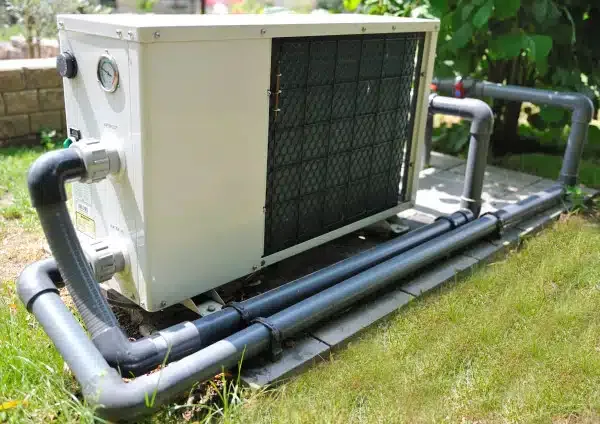 Pompe à chaleur de piscine installée sur des dalles dans le jardin, raccordée par des tuyaux PVC gris