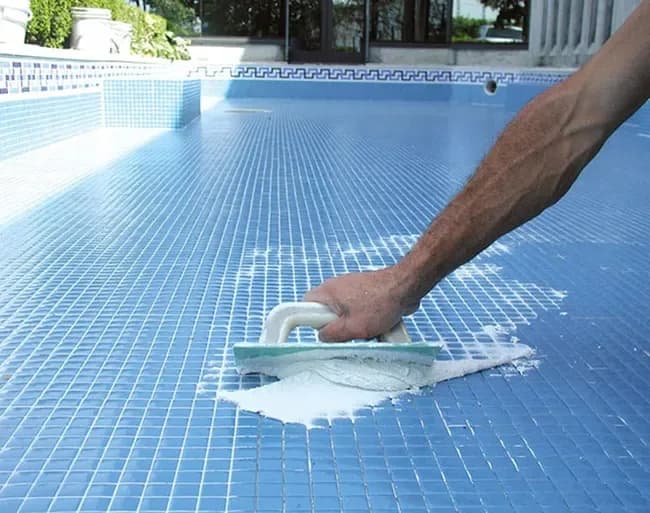 Main d’un technicien appliquant le joint blanc à la taloche sur une mosaïque bleue neuve au fond d’une piscine vide